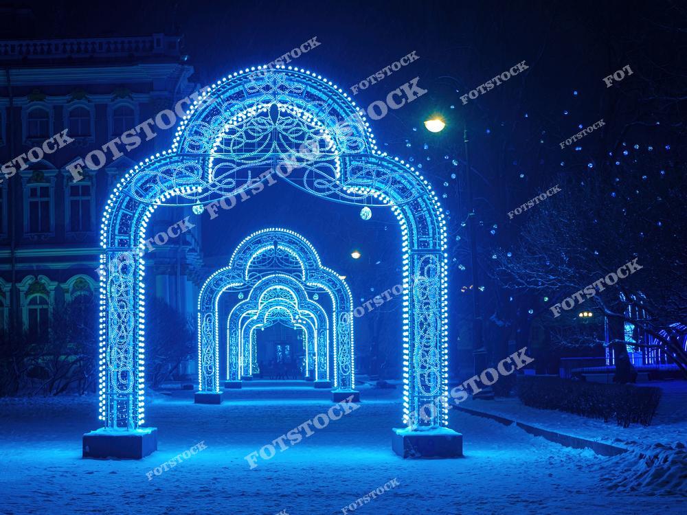 На фотографии изображены яркие светящиеся арки, установленные на одной из улиц Санкт-Петербурга в зимний период. Заснеженная дорожка подчеркивает волшебную атмосферу ночного города. Иллюминация в холодных синих тонах создает впечатление зимней сказки. Это место становится популярным среди горожан и туристов в период новогодних праздников.